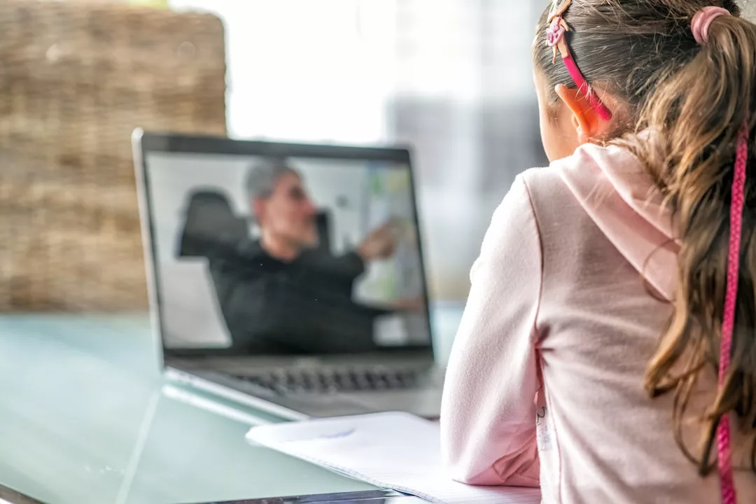 Ein junges Mädchen mit einem rosafarbenen Stirnband besucht einen Online-Kurs über effizientes Lernen und beobachtet ihren Lehrer auf dem Laptop-Bildschirm, der sein Notebook auf dem Tisch liegen hat.