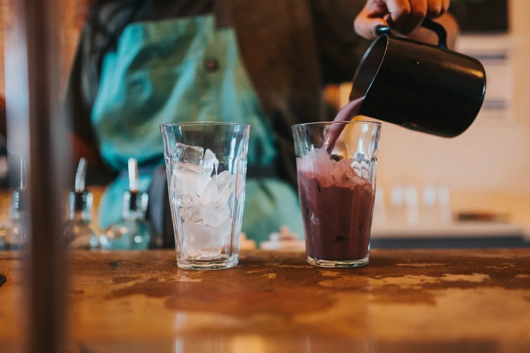 Eine Person in einer blaugrünen Schürze gießt ein lilafarbenes, gesundes Smoothies-Getränk aus einem schwarzen Krug in ein Glas mit Eis, das neben einem weiteren Glas mit Eis auf einer Theke steht.