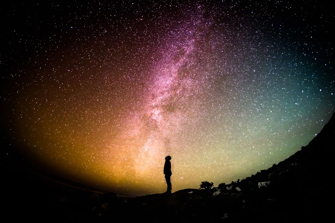 Eine Person steht als Silhouette vor einem farbenfrohen, sternenübersäten Nachthimmel, über dem die Milchstraße zu sehen ist. Sie fängt das Wunder der Astronomie ein und dient als inspirierende Einführung in dieses faszinierende Hobby.