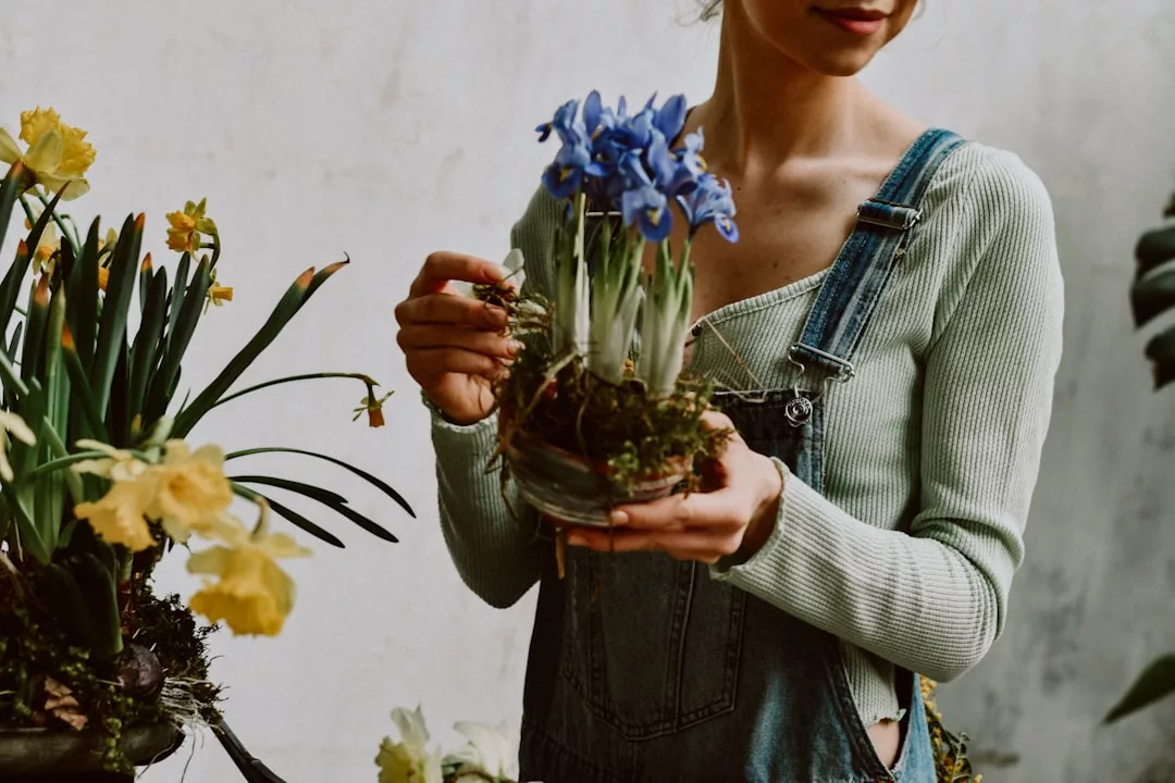 Eine Person in Jeans-Overall hält einen Glastopf mit blauen Schwertlilien, umgeben von gelben Blumen, und wirbt für Indoor-Gartenarbeit und praktische Tipps vor einem hellen Hintergrund.