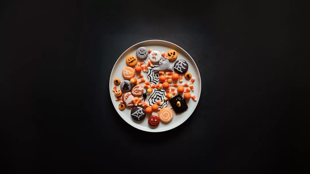 Ein weißer Teller mit verschiedenen Bonbons und Keksen zum Thema Halloween, inspiriert von der molekulare Gastronomie, verziert mit Kürbissen, Spinnennetzen und leuchtenden orangen und schwarzen Farben auf schwarzem Hintergrund.
