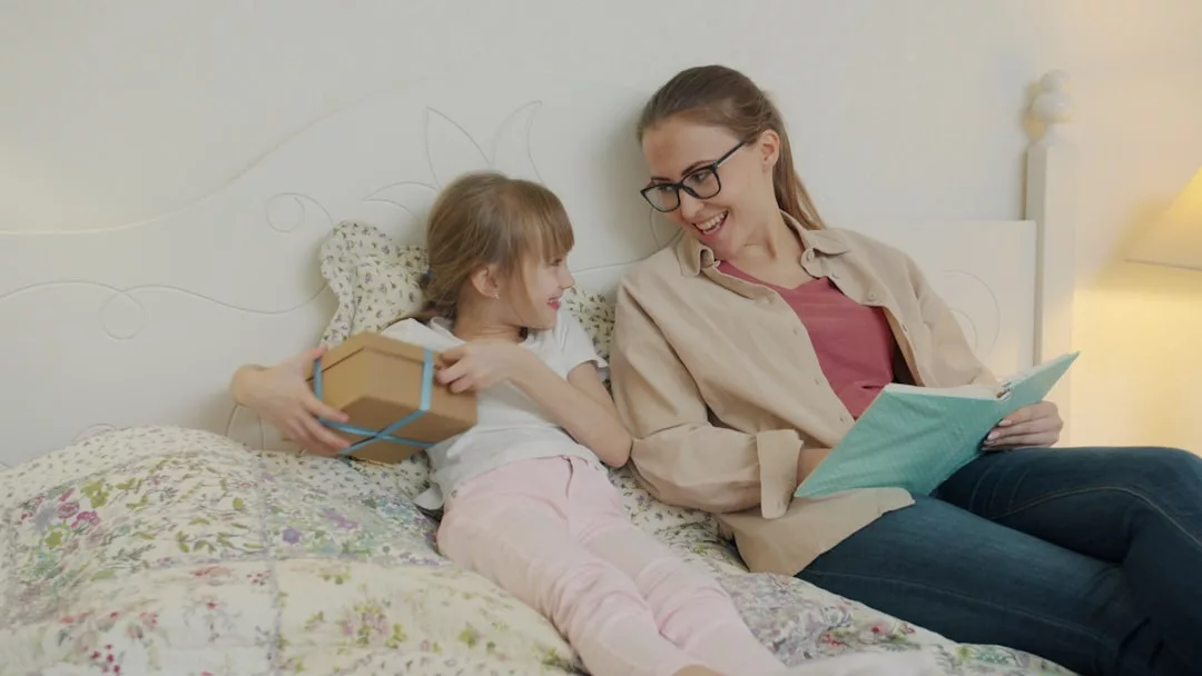 Eine Frau und ein junges Mädchen sitzen auf einem Bett und lächeln gemeinsam, während das Mädchen eine Geschenkbox und die Frau ein Dokument in der Hand hält - eine starke Bindung und eine liebevolle Eltern-Kind-Kommunikation in einem besonderen Moment.