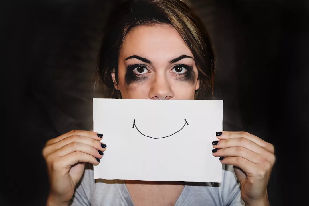Eine Person mit verschmiertem Augen-Make-up hält ein Papier mit einem gezeichneten Lächeln über ihren Mund, das ihren Gesichtsausdruck kontrastiert - ein eindrucksvolles Bild für Emotionale Intelligenz und wie wir sie entwickeln, um echten Erfolg zu finden.