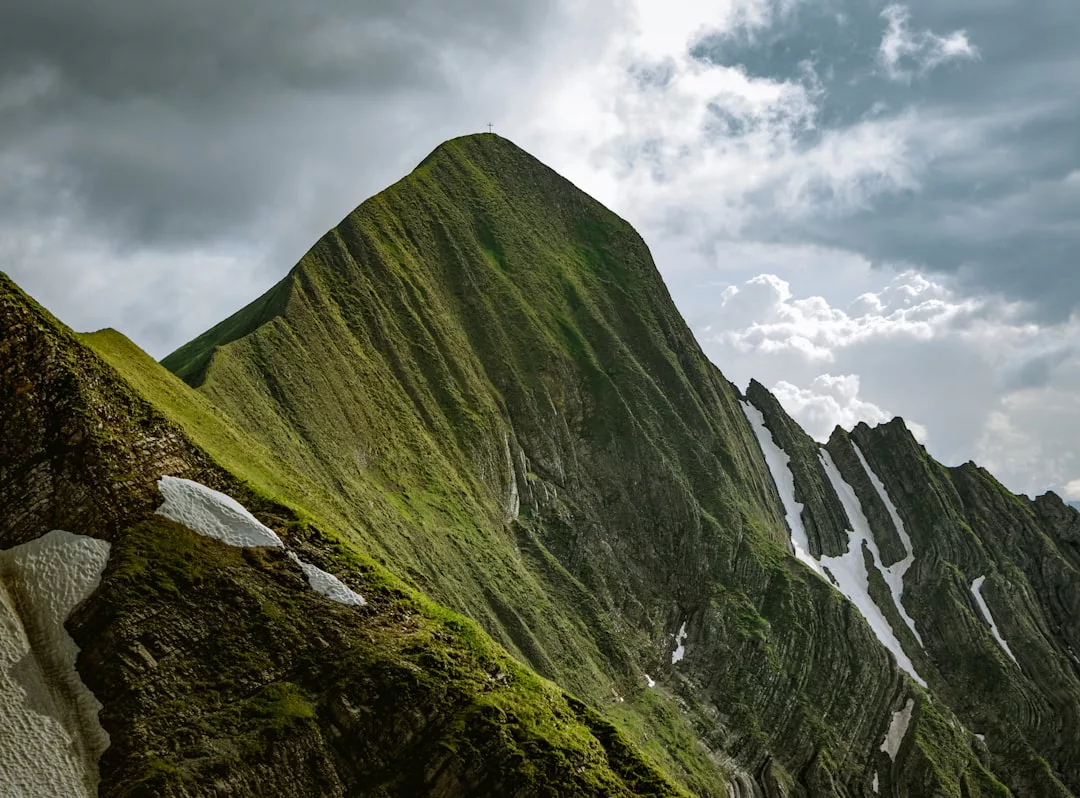 Ein steiler, grasbewachsener Berggipfel mit Schneeflecken erhebt sich unter einem wolkenverhangenen Himmel und erinnert an Emotionale Stärke und Widerstandsfähigkeit angesichts der Größe der Natur.