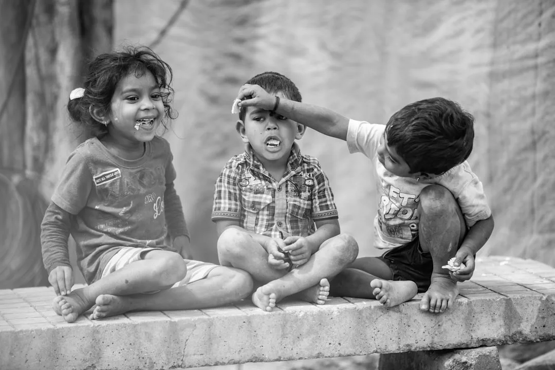 Drei kleine Kinder sitzen barfuß auf einem Felsvorsprung, wobei ein Kind spielerisch den Kopf eines anderen berührt, während das dritte in die Kamera lächelt - ein authentischer Moment, der die Kunst der Empathie in diesem Schwarz-Weiß-Bild einfängt.