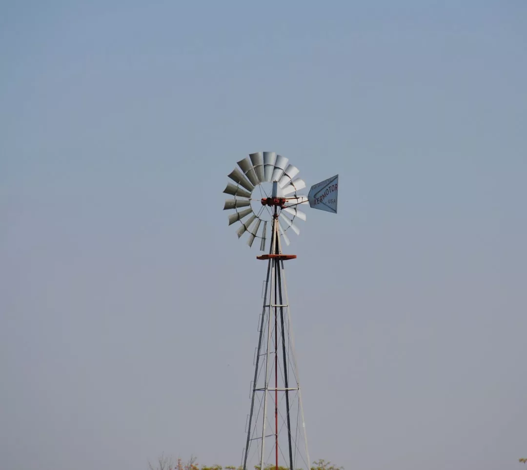 Ein Metallwindrad mit rotierendem Flügel und Schwanz steht vor einem klaren blauen Himmel und zeigt effiziente Methoden der Energieerzeugung, wobei am unteren Bildrand eine minimale Vegetation sichtbar ist.