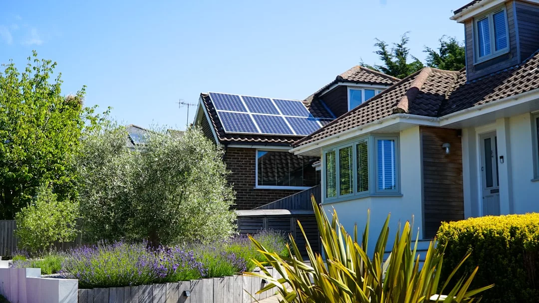 Ein Haus mit Ziegeldach und weißer Fassade steht neben einem anderen Haushalt, in dem Sonnenkollektoren den Energieverbrauch reduzieren. Der angelegte Garten im Vordergrund ist mit Sträuchern und Bäumen geschmückt.