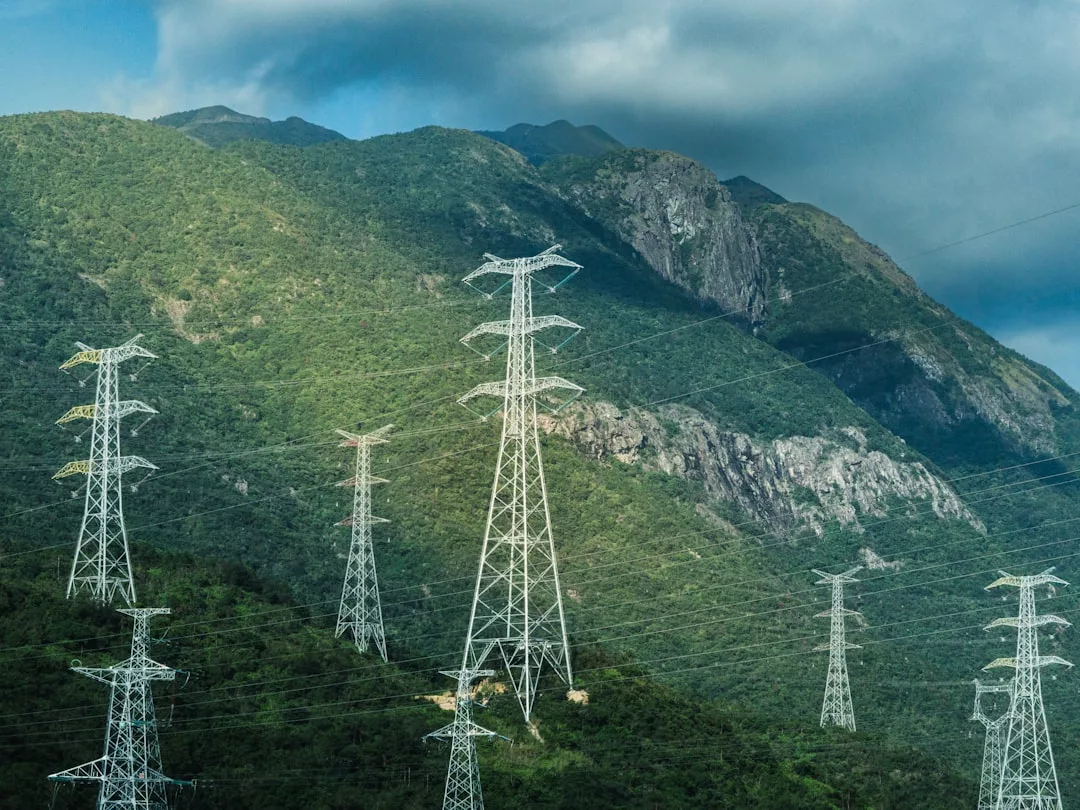 Hochspannungsmasten symbolisieren die moderne Energieversorgung und erstrecken sich über eine grüne, bergige Landschaft unter einem teilweise bewölkten Himmel.