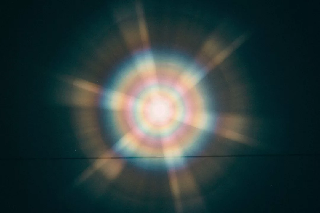 Ein kreisförmiges, mehrfarbiges Lichtmuster mit strahlenden Strahlen erzeugt eine hypnotisierende Aura vor einem dunklen Hintergrund, die an eine Aura-Welt erinnert.