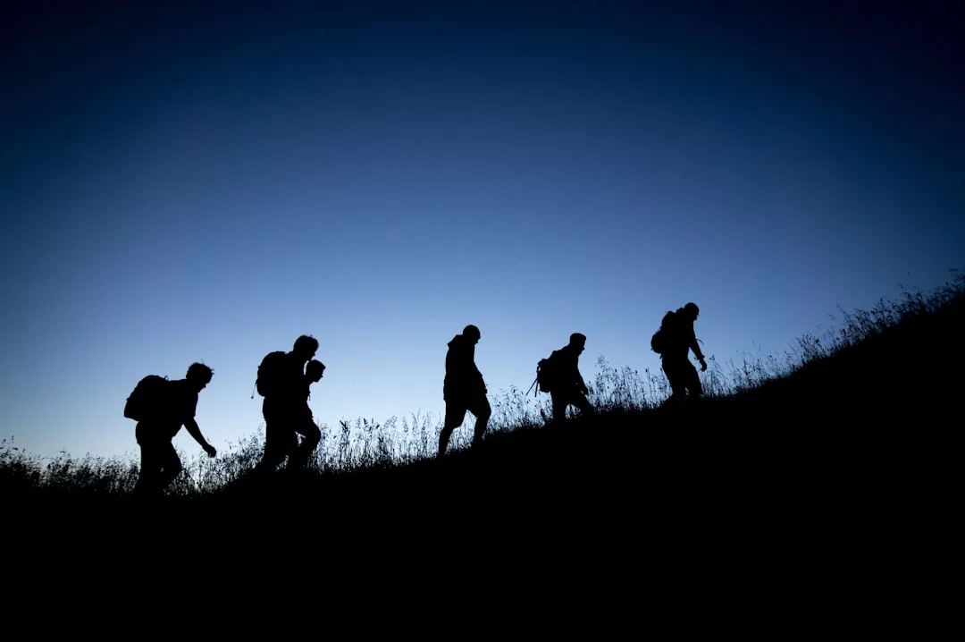 Fünf Personen mit Rucksäcken wandern in einer Reihe entlang eines grasbewachsenen Hangs bergauf, als Silhouette vor einem klaren blauen Himmel - ein Symbol für Führung und Teamarbeit auf dem Weg der Entwicklung.
