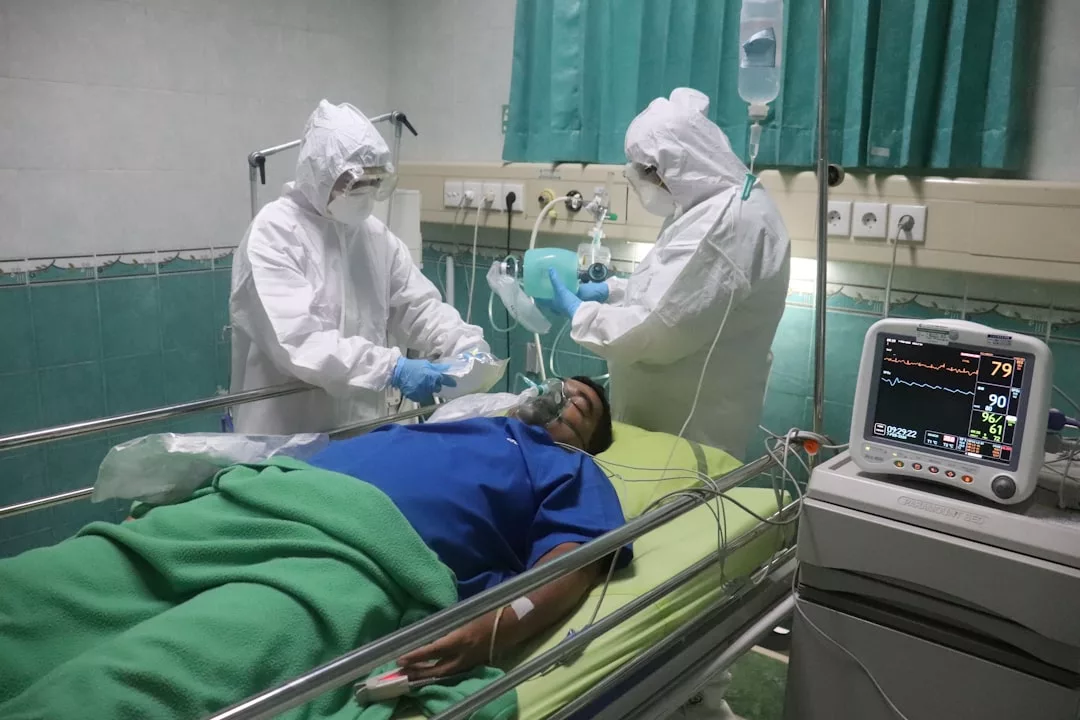 Zwei Mitarbeiter des Gesundheitswesens in Schutzkleidung kümmern sich um einen Patienten, der mit einer Sauerstoffmaske auf einem Krankenhausbett liegt, neben einem Überwachungsgerät, das die Vitalwerte anzeigt, und demonstrieren so erfolgreiche Teamarbeit und Engagement.