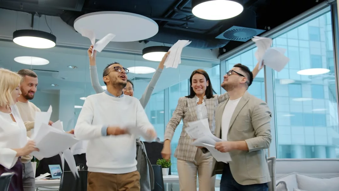 Eine Gruppe von Kollegen in einem Büro feiert ihren Projekterfolg, indem sie Papiere in die Luft werfen, lächeln und an Schreibtischen mit großen Fenstern im Hintergrund stehen.