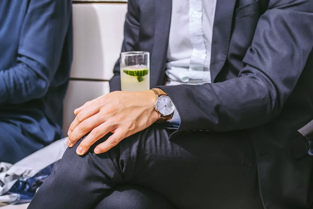 Bewerber in dunklem Anzug sitzt mit überschlagenen Beinen, hält ein Getränk mit Eis und Gurkenscheibe in einer Hand und trägt eine silberne Armbanduhr - ein stilvoller Auftritt fürs Vorstellungsgespräch.