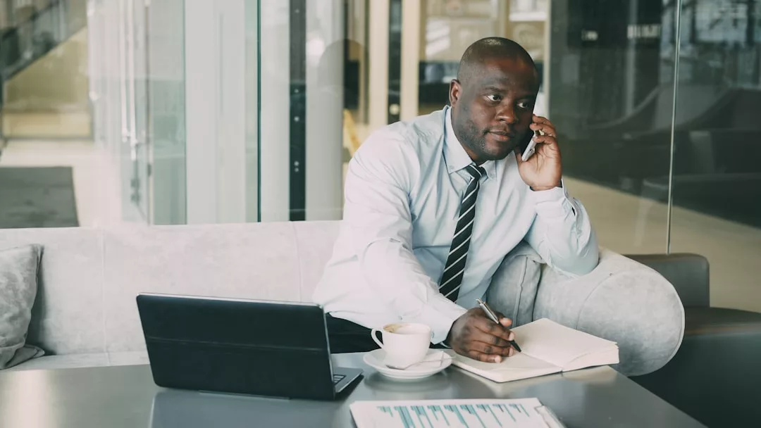 Ein Mann in Business-Kleidung sitzt auf einem Sofa, telefoniert und schreibt in ein Notizbuch - er balanciert Laptop, Dokumente und Kaffee - während er seine Erfolgsstrategien für den beruflichen Aufstieg plant.