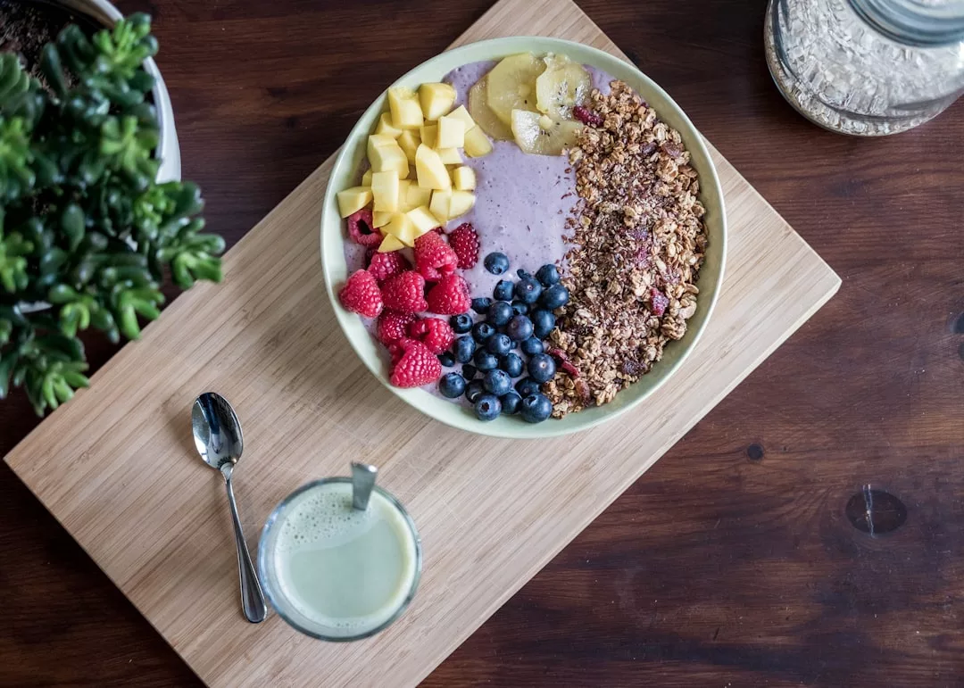 Eine Smoothie-Bowl mit Müsli, Blaubeeren, Himbeeren, Mangowürfeln und Bananenscheiben steht auf einem Holzbrett - perfekt für leckere Ernährungstipps Kinder und eine ausgewogene Ernährung im Kleinkindalter.