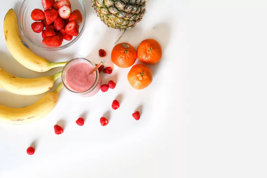 Bananen, Erdbeeren, eine Ananas, Mandarinen, Himbeeren und ein Glas rosa Smoothie sind auf einer weißen Fläche angeordnet - perfekt für alle, die Wert auf gesunde Ernährung und Fitness legen.