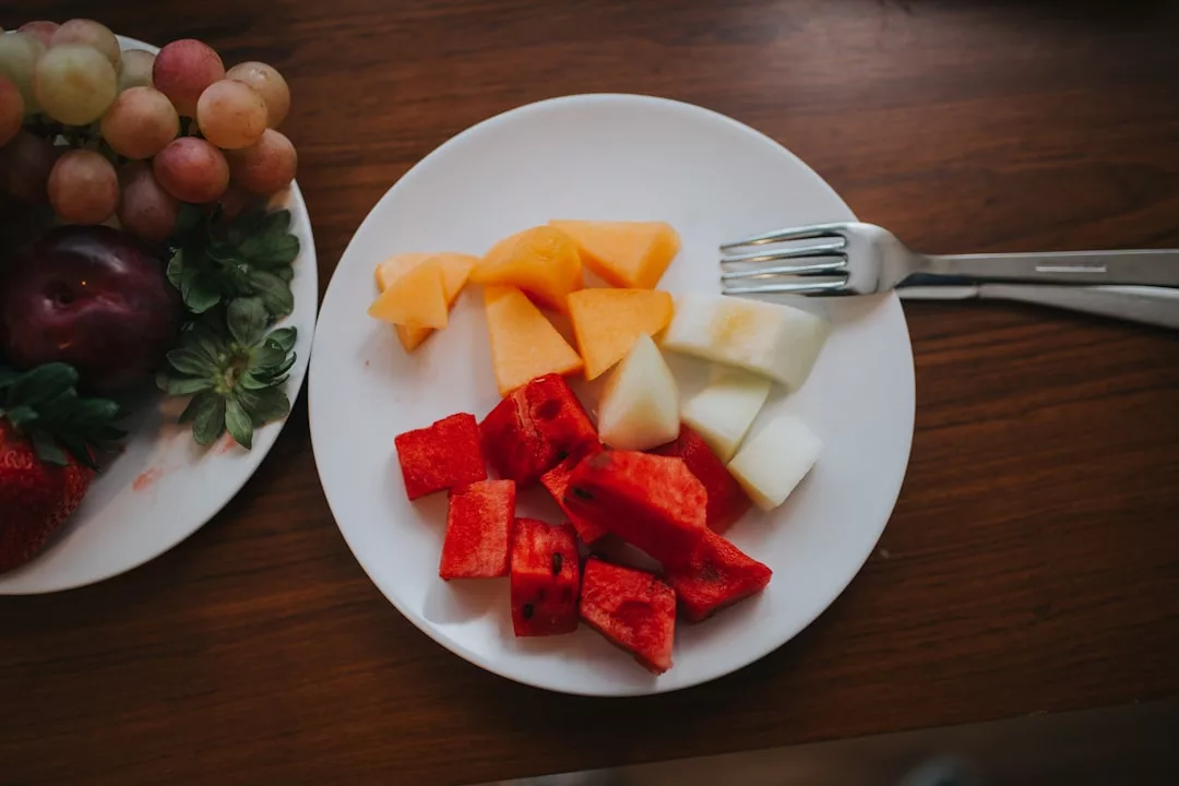 Ein weißer Teller mit verschiedenen Obststücken, darunter Melone, Honigmelone und Wassermelone, wirbt neben einer Gabel auf einem Holztisch für gesunde Ernährung. Ein weiterer Teller mit Weintrauben und Erdbeeren ist teilweise sichtbar.