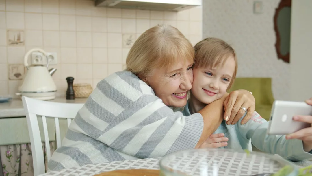 Eine ältere Frau und ein junges Mädchen umarmen sich und lächeln, während sie gemeinsam ein Selfie am Küchentisch machen, um einen Moment der Bindung und Freude in ihren Familienbeziehungen festzuhalten.