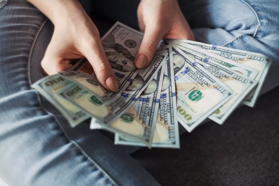 Eine Person, die mehrere US-Hundertdollarscheine in der Hand hält und auffächert, während sie in Jeans auf dem Boden sitzt, symbolisiert finanziellen Erfolg und den Traum von finanzieller Freiheit.