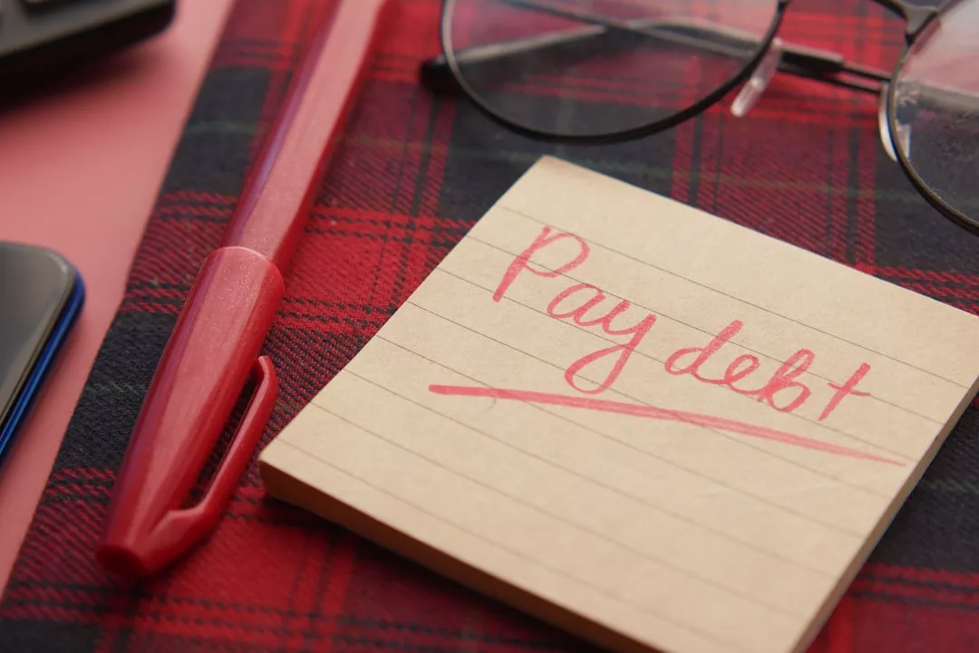 Auf einer rot karierten Fläche liegen ein roter Stift, eine Brille und ein Notizblock mit der Aufschrift "Schulden bezahlen" - die perfekten Werkzeuge für deine finanzielle Zielsetzung und Geldplanung.