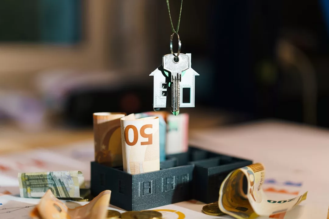 Ein metallischer Schlüsselanhänger in Form eines Hauses mit einem Schlüssel hängt über gerollten Euro-Banknoten in einem kleinen schwarzen Tablett, das die Immobilienfinanzierung symbolisiert, während weiteres Bargeld auf dem Tisch verstreut ist.