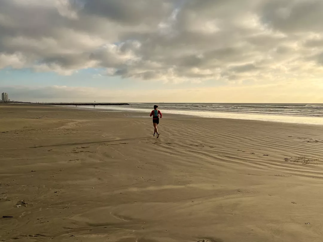 Ein fitter Mensch joggt allein an einem breiten Sandstrand unter einem wolkenverhangenen Himmel, mit dem Meer und entfernten Gebäuden im Hintergrund, und genießt einen entspannten Moment, der sich wie ein echter Fitnessurlaub anfühlt.
