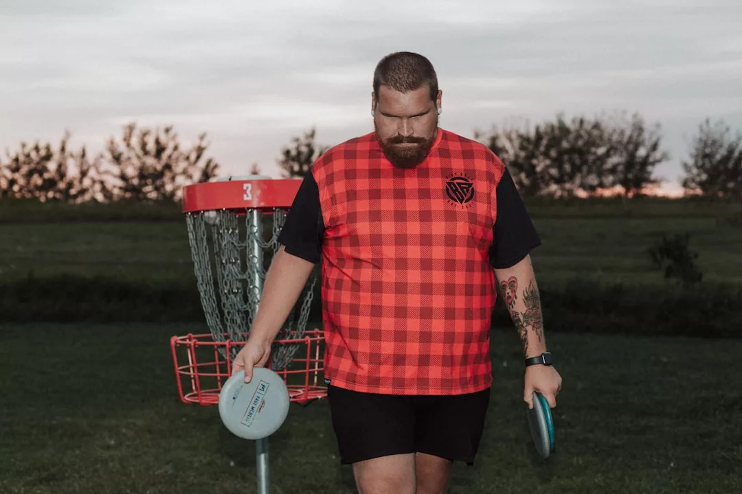 Ein Mann in einem rotkarierten Hemd hält zwei Scheiben, während er neben einem Disc-Golf-Korb auf einer Wiese in der Abenddämmerung steht und den Sport genießt, der perfekt für Fitness-Enthusiasten und alle ist, die einen fitten und gesunden Lebensstil suchen.