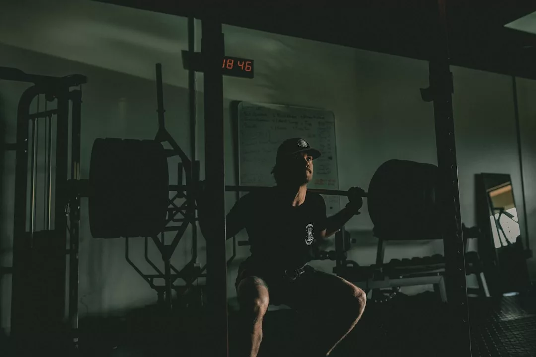 Eine Person mit Kappe führt eine Kniebeuge mit einer schwer belasteten Langhantel in einem schwach beleuchteten Fitnessstudio durch, das effektive Übungen und Fitnesstraining für Männer vorstellt. Im Hintergrund sind eine Digitaluhr und eine Tafel zu sehen.