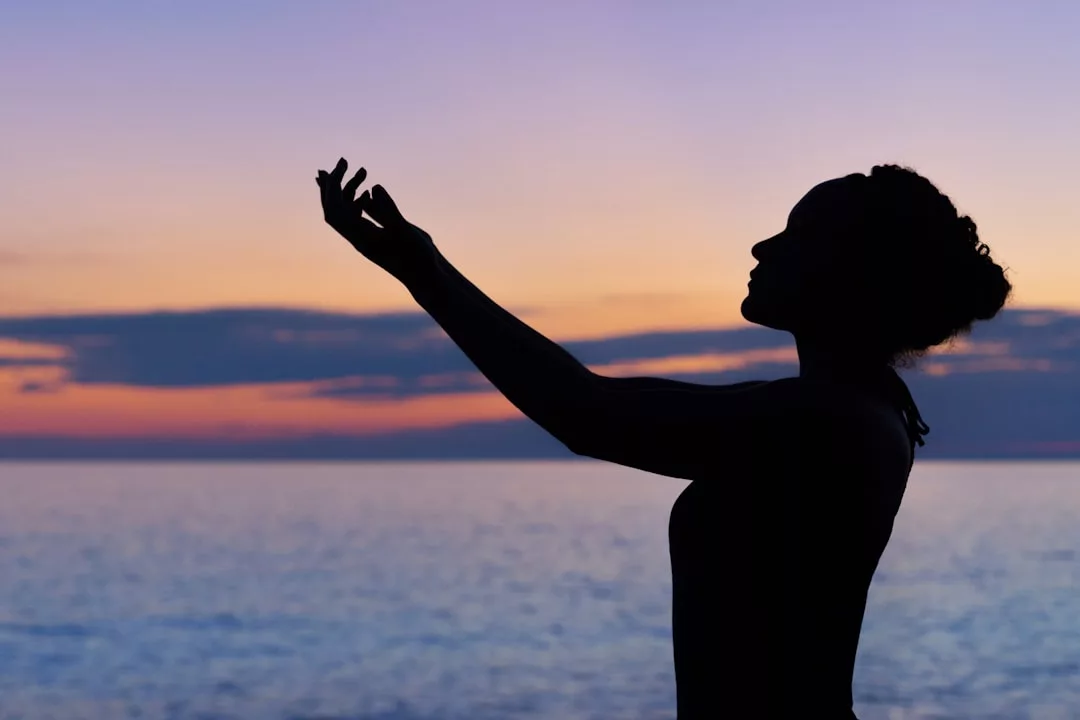 Silhouette einer Person mit erhobenem Arm, die bei Sonnenuntergang am Meer steht, während der bunte Himmel in der ruhigen Atmosphäre Schwingungen und ein Gefühl von Frequenzheilung hervorruft.