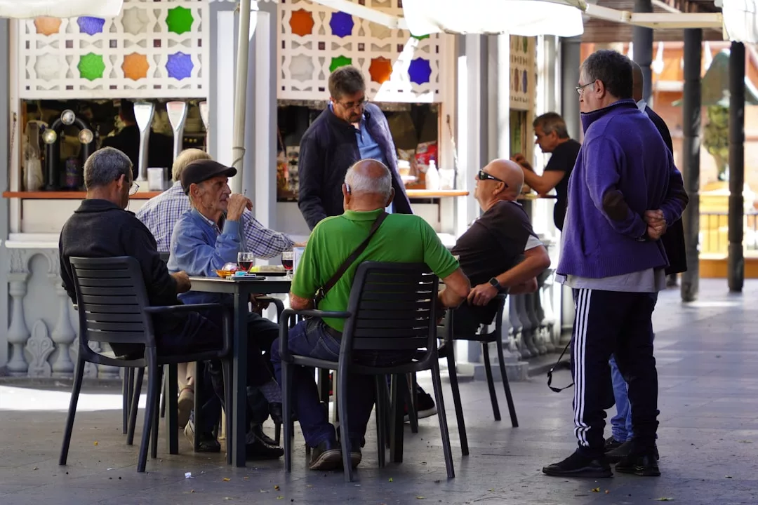 Eine Gruppe älterer Männer sitzt an einem Cafétisch im Freien und unterhält sich bei Getränken und Snacks, während sie Freundschaft pflegen, während zwei andere in der Nähe stehen.