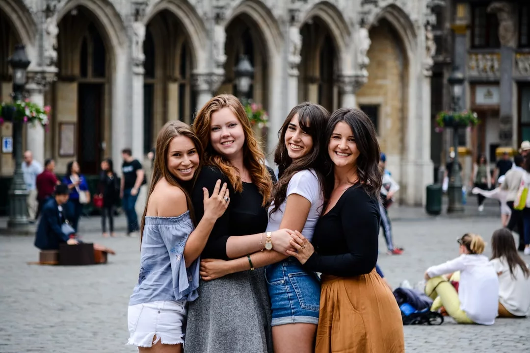 Vier junge Frauen stehen dicht beieinander und lächeln in die Kamera auf einem historischen europäischen Stadtplatz, mit gewölbten Gebäuden und Menschen im Hintergrund - ein schönes Beispiel dafür, wie man Freundschaften im Erwachsenenalter pflegen kann.