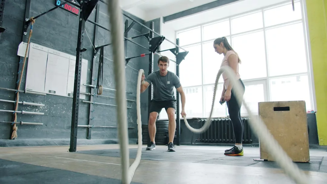 Ein Mann betreibt funktionelles Training mit Kampfseilen in einem Fitnessstudio, während eine Frau in der Nähe steht und diesem effektiven Workout für den ganzen Körper zusieht.