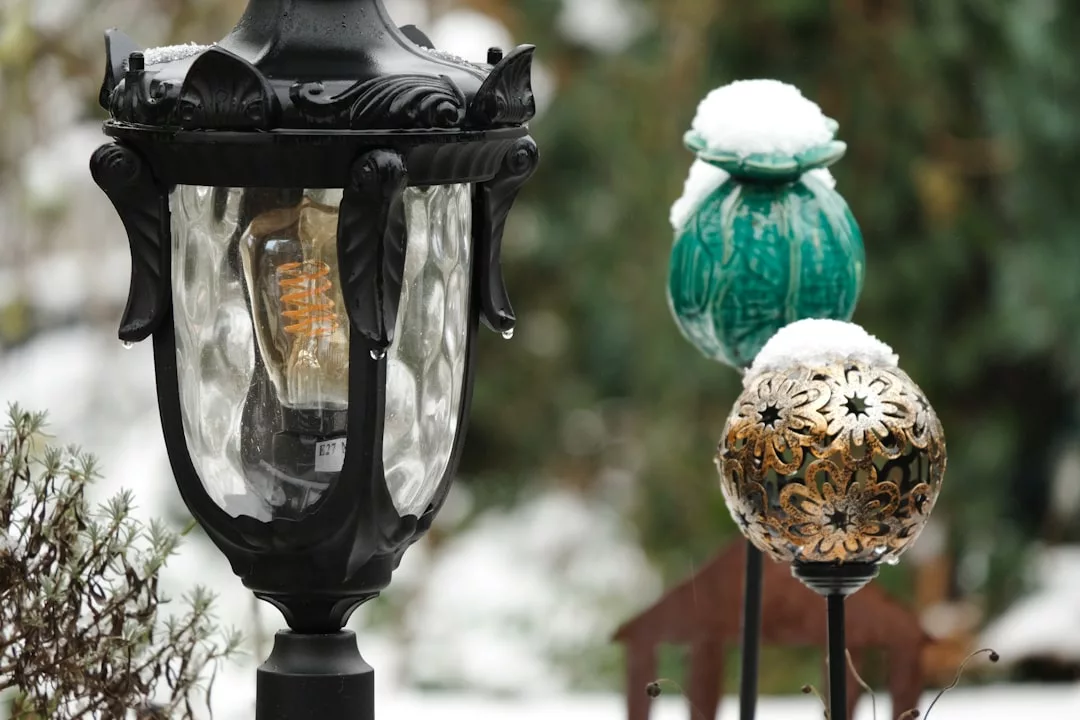 Nahaufnahme einer schwarzen Außenlaterne und zweier dekorativer Gartenornamente, die mit Schnee bedeckt sind und stilvolle Akzente zur winterlichen Gartendekoration im Außenbereich setzen, vor einem unscharfen Hintergrund aus Grünzeug und Schnee.