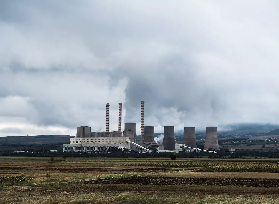Ein großes Industriekraftwerk mit mehreren Schornsteinen und Kühltürmen stößt unter bewölktem Himmel Rauch aus. Es liegt in einer flachen, ländlichen Landschaft und verdeutlicht die Notwendigkeit einer Umstellung auf nachhaltige Energiequellen wie Geothermie.