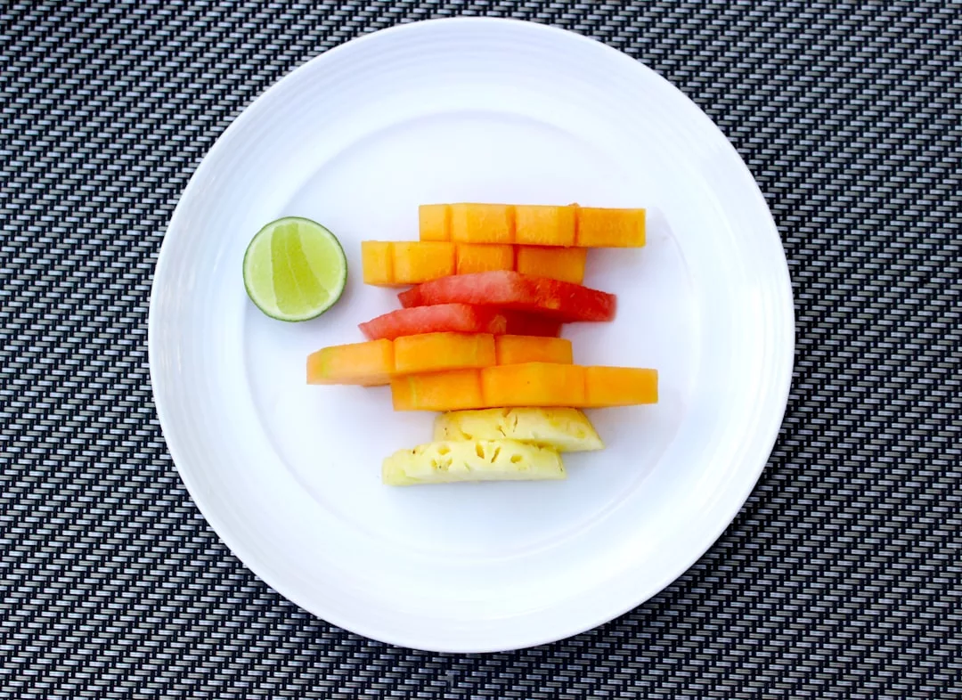 Ein weißer Teller mit fein säuberlich angeordneten Papaya-, Wassermelonen- und Ananasscheiben sowie einer halben Limette auf einem gewebten dunklen Tischset - perfekt für eine gesunde Kinderernährung.