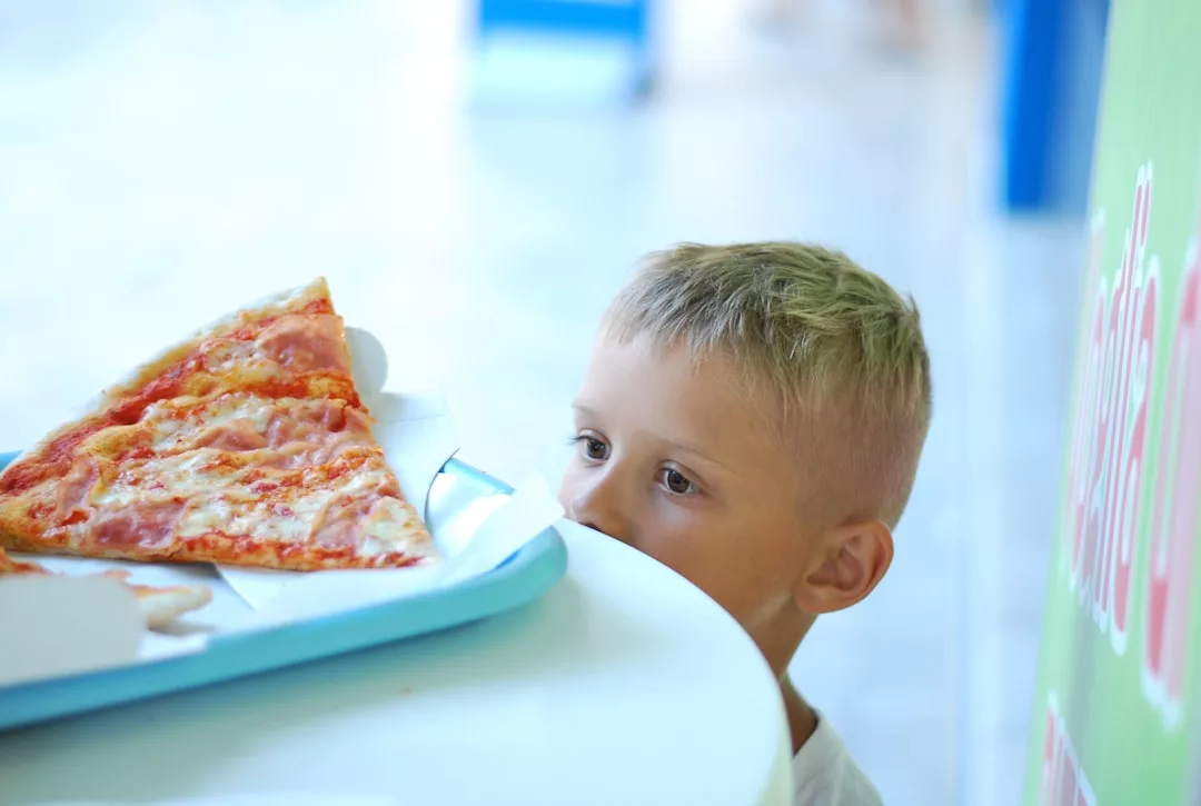 Ein kleiner Junge blickt über einen Tisch auf ein großes Stück Pizza auf einem blauen Tablett in einem hell erleuchteten Raum und erinnert uns daran, wie wichtig gesunde Kinderernährung ist - hier sind praktische Tipps für Eltern, um ausgewogene Mahlzeiten schmackhaft zu machen.