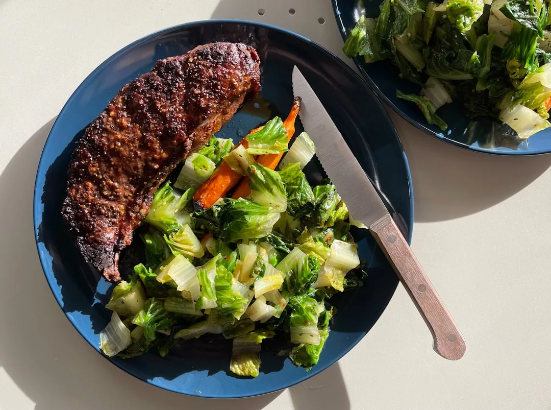 Ein blauer Teller mit einem gebratenen Steak und gebratenem Gemüse, darunter Karotten und Grünzeug, ist ein Beispiel für gesundes Kochen. Ein Messer liegt neben dem Essen, und ein Beilagenteller mit weiterem Gemüse ist teilweise sichtbar - ideal für alle, die preisgünstige Essensideen suchen.