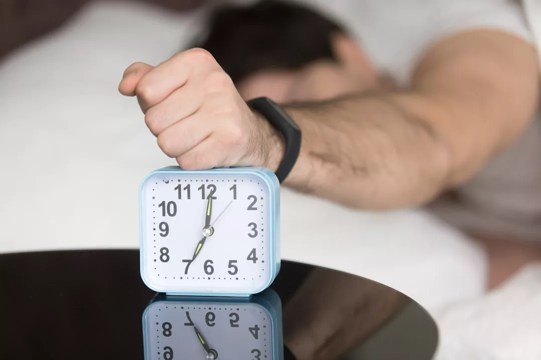 Eine im Bett liegende Person streckt einen Arm aus, um die Spitze eines blauen Weckers zu treffen, der 7:00 Uhr auf einem Nachttisch anzeigt - der perfekte Start für gesunde Schlafgewohnheiten und erholsame Nächte.