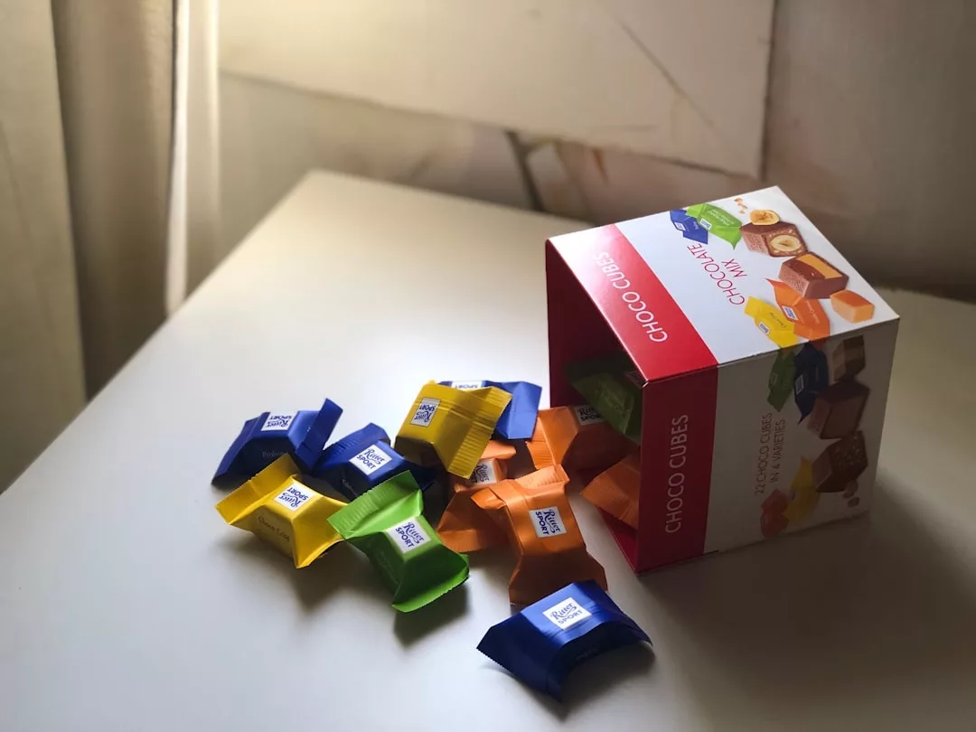 Eine Schachtel mit der Aufschrift "Choco Cubes" steht auf einem weißen Tisch und verteilt nahrhafte Snacks - einzeln verpackte Pralinen in gelber, grüner, blauer und oranger Verpackung - ideal als Schulsnacks für Kinder.
