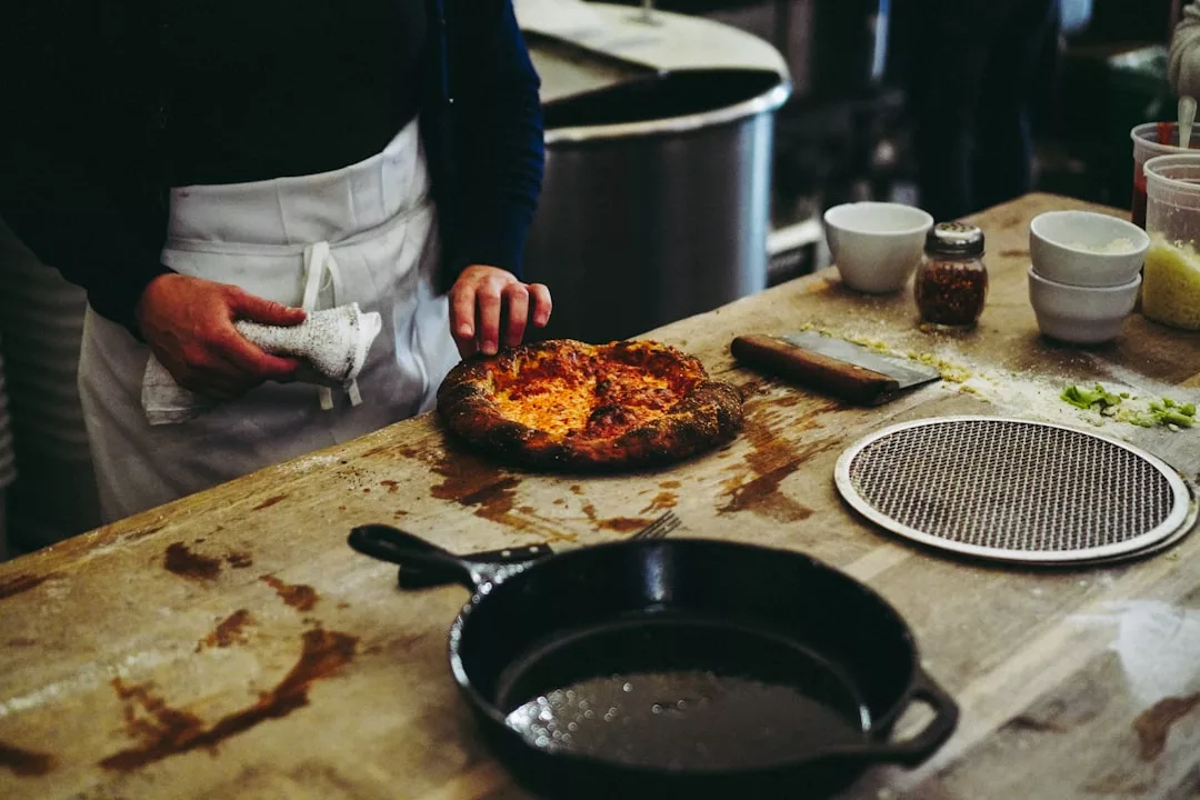Eine Person in Schürze steht an einer Holztheke mit einer gebackenen Pizza, einer gusseisernen Pfanne und mehreren Schüsseln und präsentiert die feine Küche in einer einladenden Umgebung.