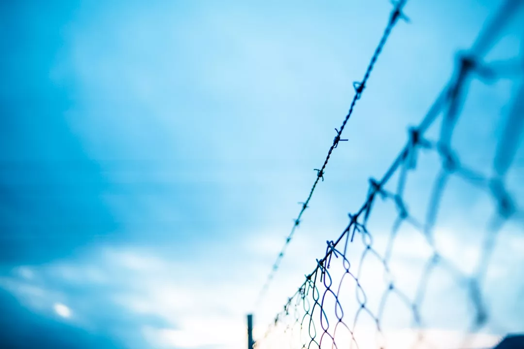 Nahaufnahme eines Stacheldrahtzauns vor einem blauen Himmel mit vereinzelten Wolken, die Abgrenzung und den Akt des Grenzen-Setzens symbolisieren.