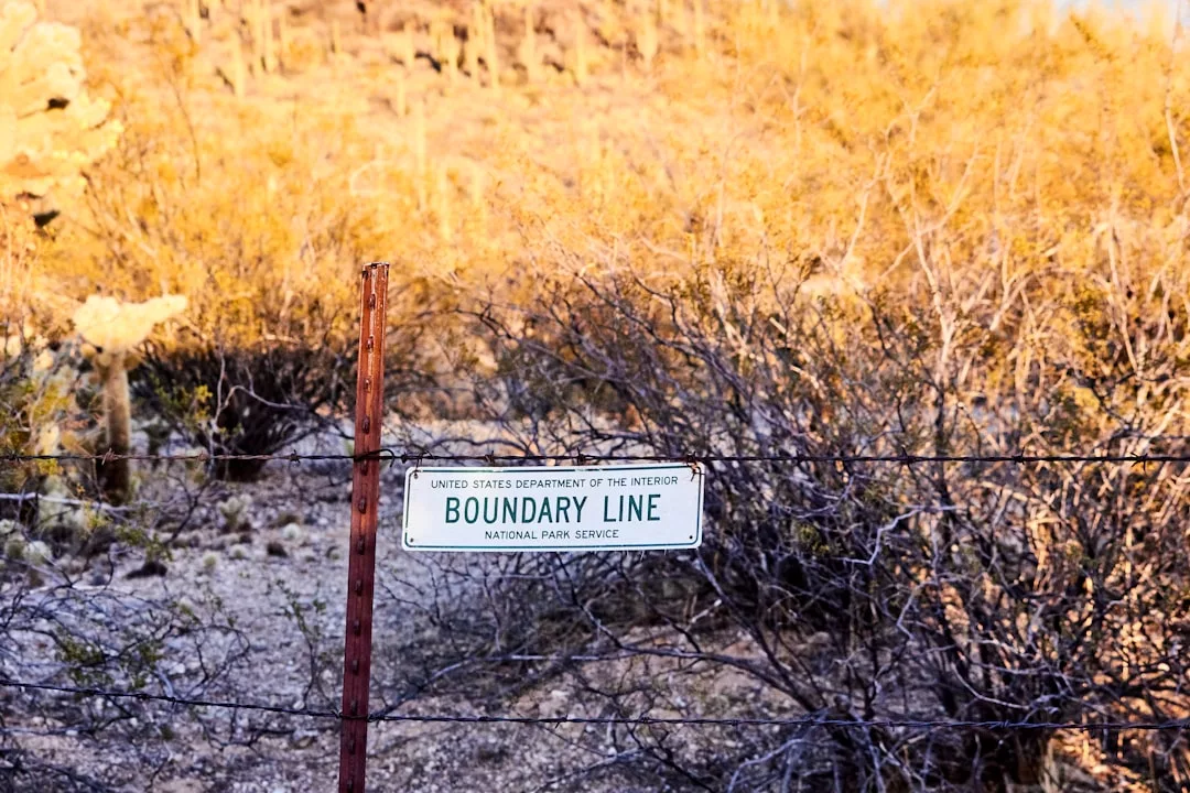 Ein Metallschild mit der Aufschrift "Boundary Line National Park Service" ist an einem Stacheldrahtzaun in einem trockenen, buschigen Gebiet angebracht und symbolisiert die Bedeutung von Grenzen setzen und Selbstbehauptung beim Schutz von Naturräumen.