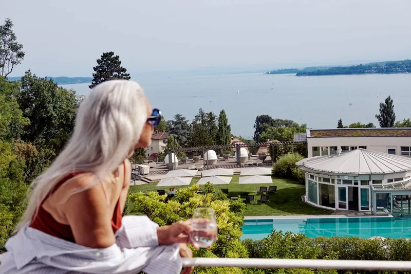 Frau mit Wasserglas auf einer Terrasse von Buchinger Wilhelmi, darunter Garten, Pool und der See