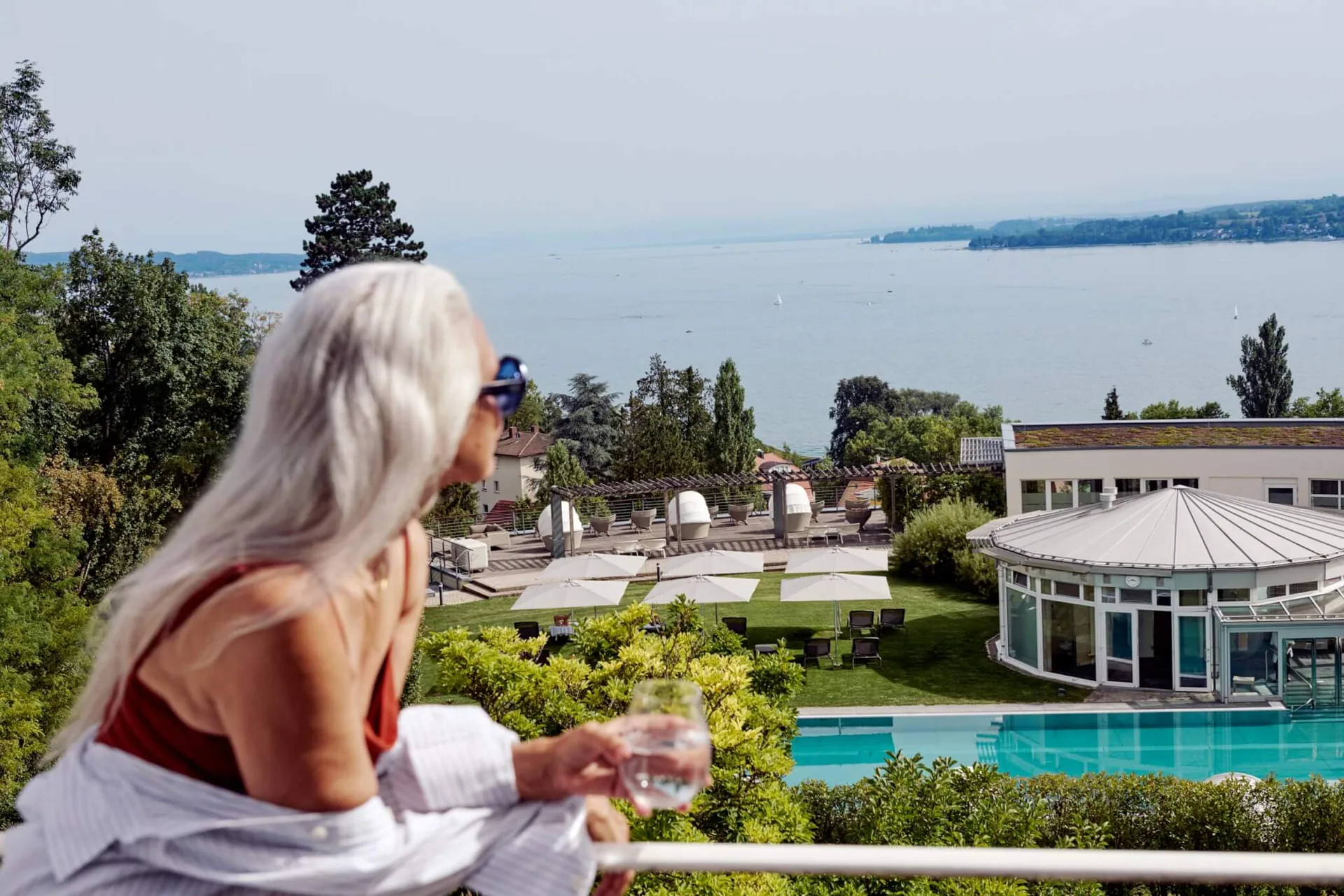 Frau mit Wasserglas auf einer Terrasse von Buchinger Wilhelmi, darunter Garten, Pool und der See