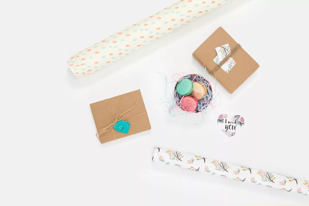 Zwei eingepackte Geschenke, eine Schale Macarons, ein herzförmiger Dankeschön-Anhänger und zwei Rollen dekoratives Geschenkpapier sind auf einer weißen Fläche arrangiert - die perfekte Inspiration für DIY-Projekte oder kreative Tipps rund ums Verpacken.