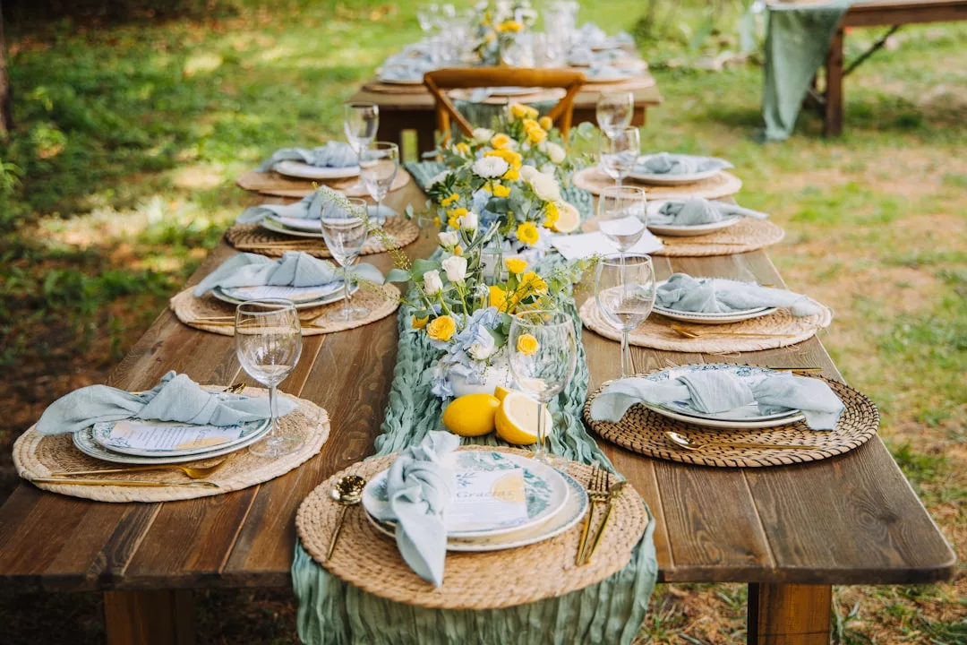 Ein rustikaler Tisch im Freien ist mit geflochtenen Tischsets, Tellern, Servietten, Gläsern, Besteck und einem floralen Mittelstück gedeckt - perfekt für Inspirationen zur Gartengestaltung und Tipps zur Gartenplanung.