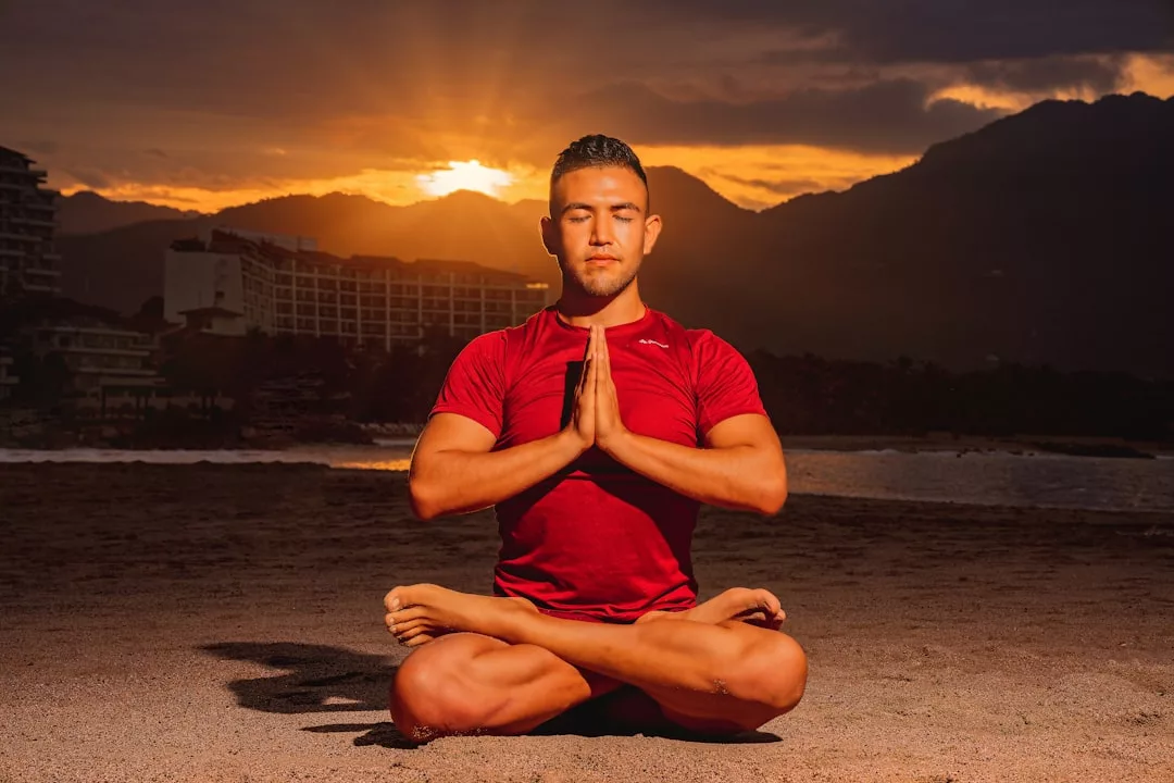 Ein Mann in einem roten Hemd praktiziert Yoga und Meditation im Lotussitz an einem Strand bei Sonnenuntergang, die Hände in Gebetshaltung und die Augen geschlossen.