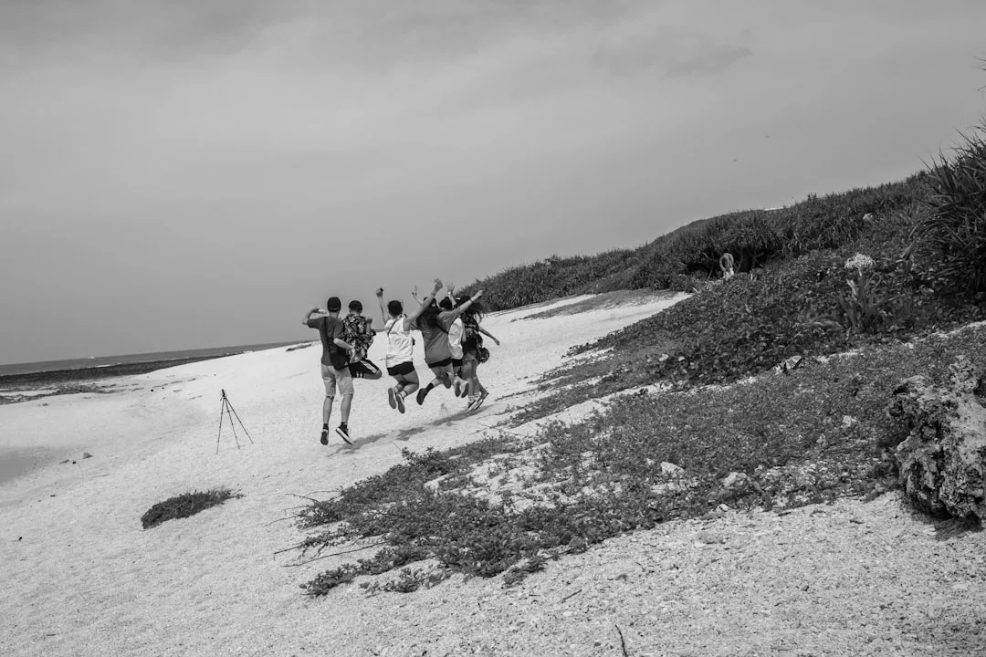 Fünf Personen springen an einem felsigen Strand in die Luft, während ein Stativ in der Nähe aufgebaut ist. Dieser fröhliche Moment wurde in Schwarz-Weiß mit Vegetation im Hintergrund aufgenommen und ist perfekt für alle, die Tipps für stressfreies Reisen mit Kindern suchen.