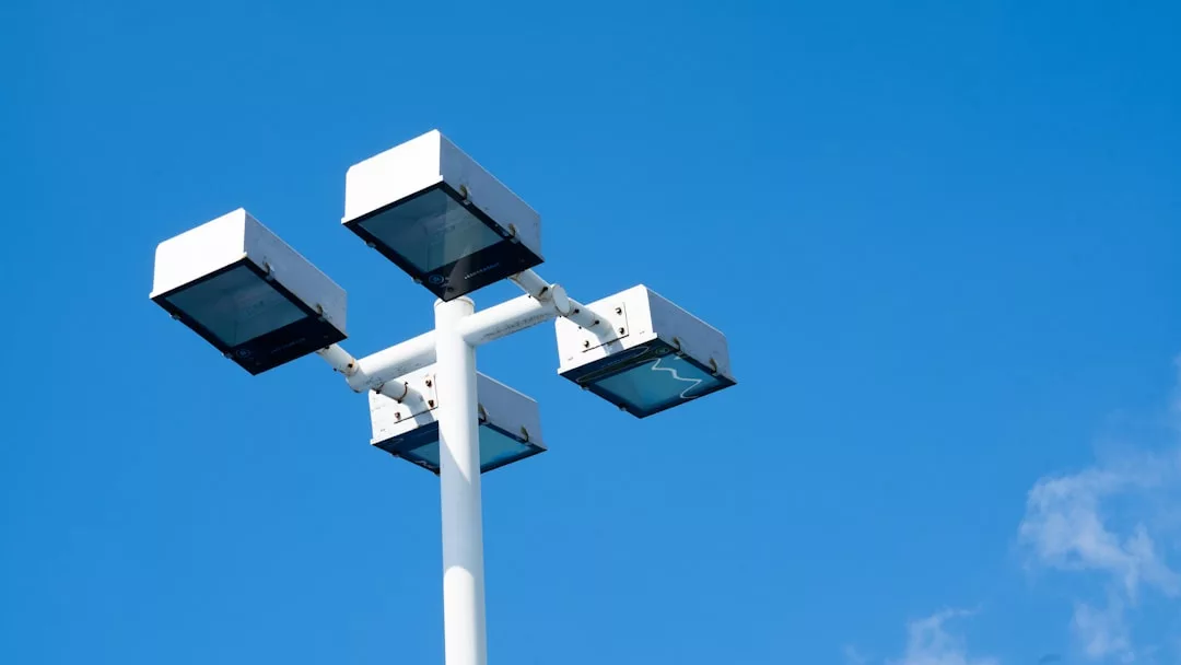 Ein weißer Mast mit vier rechteckigen Scheinwerfern ragt vor einem klaren blauen Himmel auf und stellt eine moderne Installation dar, die sich perfekt für Beleuchtungslösungen im Freien eignet.