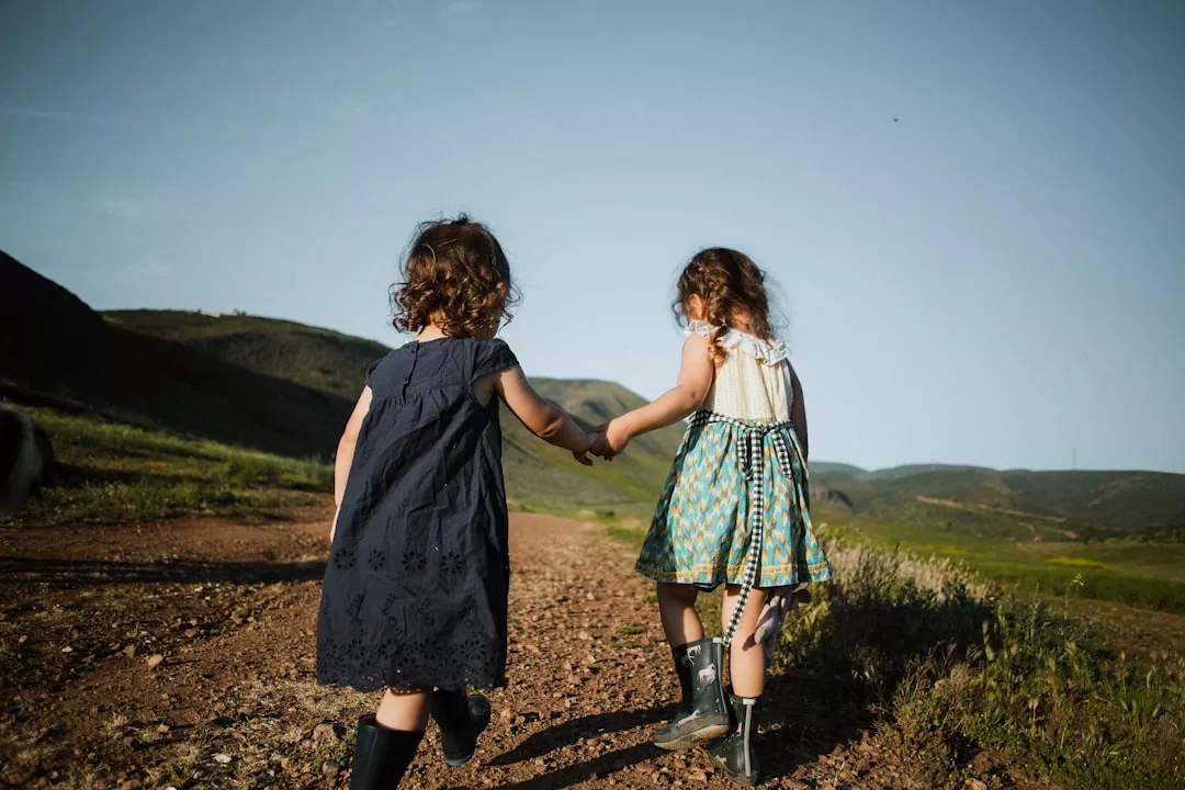 Zwei junge Mädchen in Kleidern und Stiefeln gehen Hand in Hand einen unbefestigten Weg hinunter, ihre Neugier spiegelt den Geist von Kinder für Wissenschaft und Technik begeistern wider, der sich in einer friedlichen ländlichen Landschaft zwischen Hügeln abspielt.